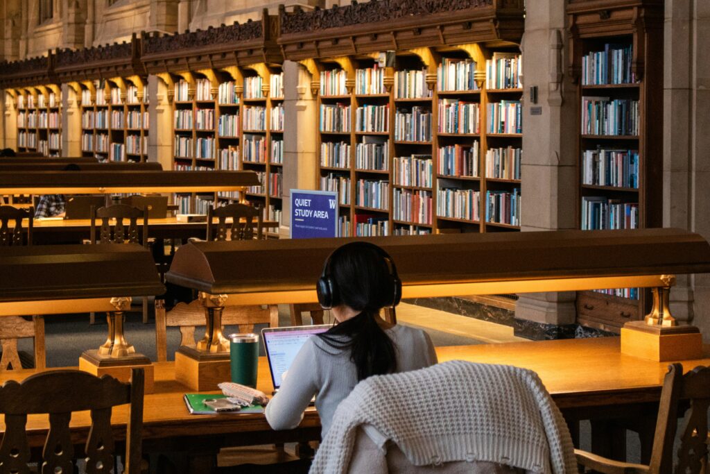 Student at a library desk with a laptop and notebooks in a quiet study area, surrounded by bookshelves.